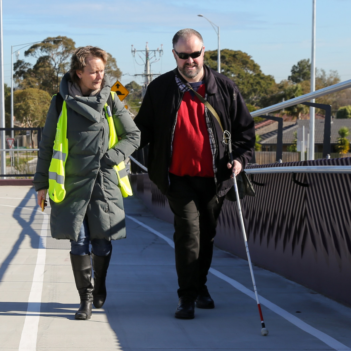 A woman in a hi-vis vest walks with a blind man using a cane.